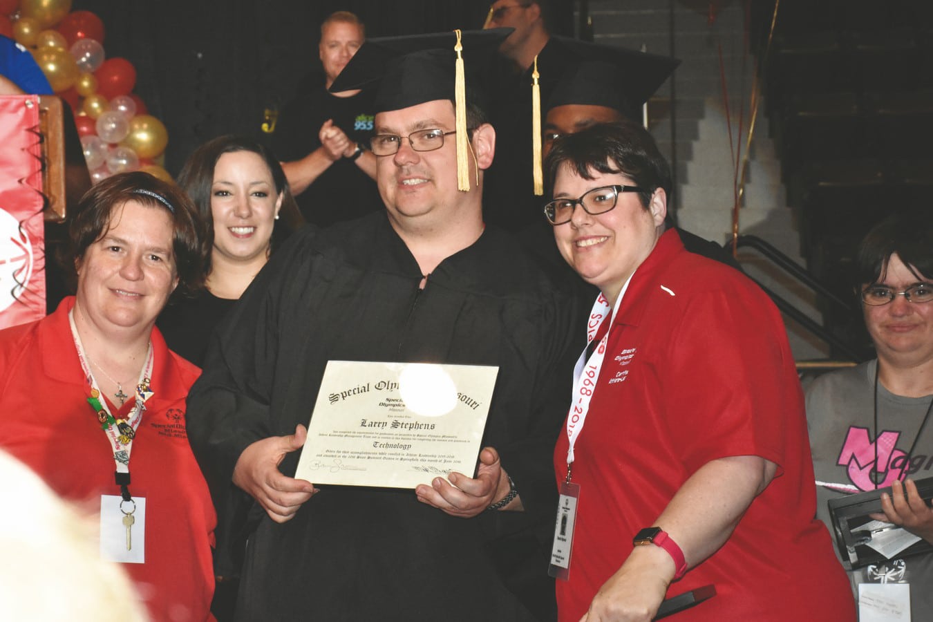 An athlete-leader in a cap and gown holds their SOMO Athlete Leadership diploma while flanked by their mentor on one side and a friend on the other