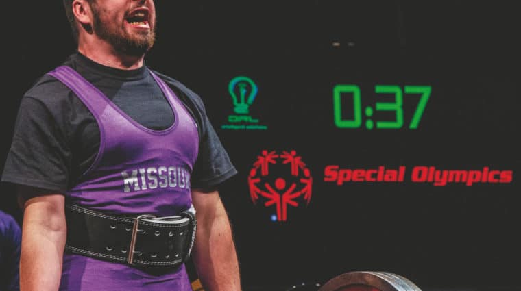 An athlete wearing a purple Team Missouri uniform successfully lifts a large amount of weight with a Special Olympics logo and countdown timer in the background