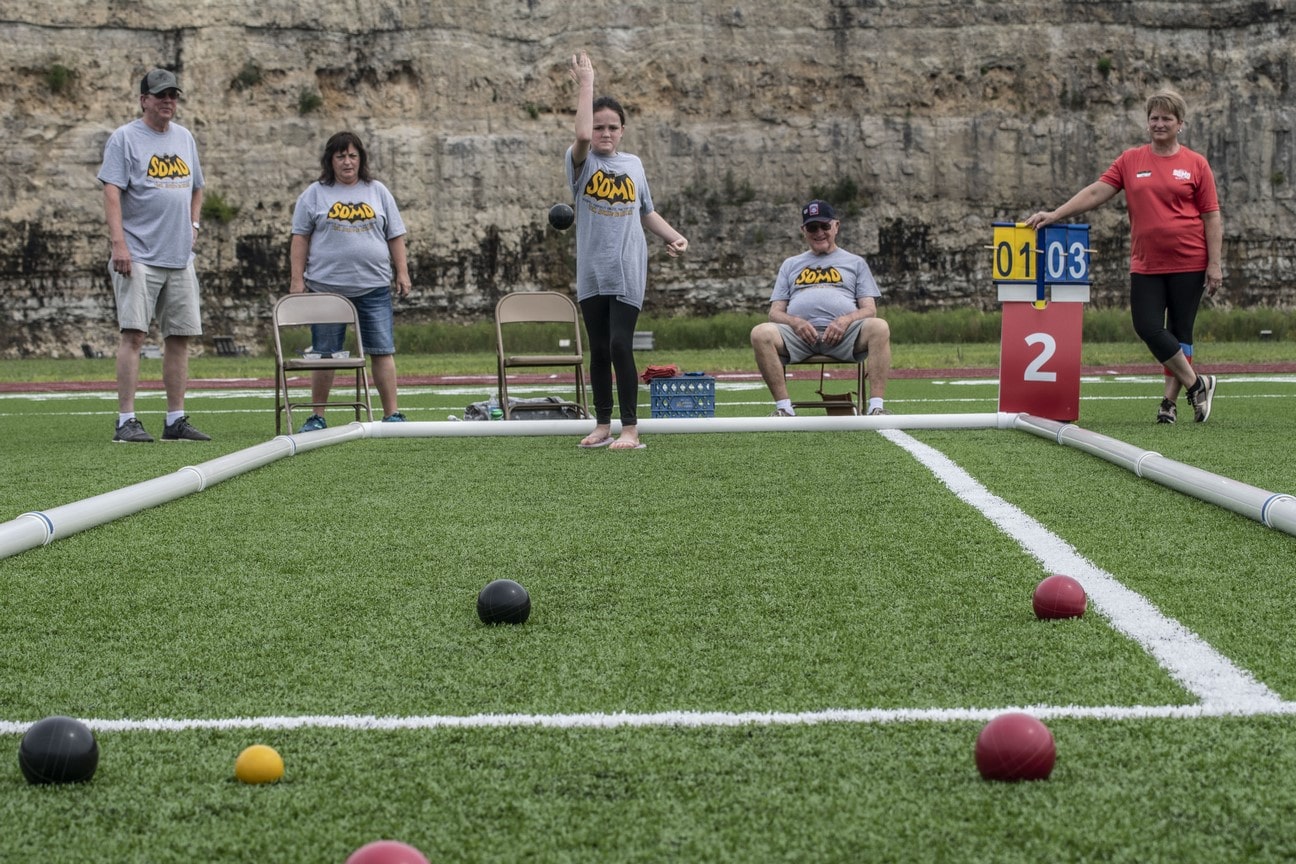 A Young Athlete releases a bocce ball from their hand to roll down the turf field toward the camera
