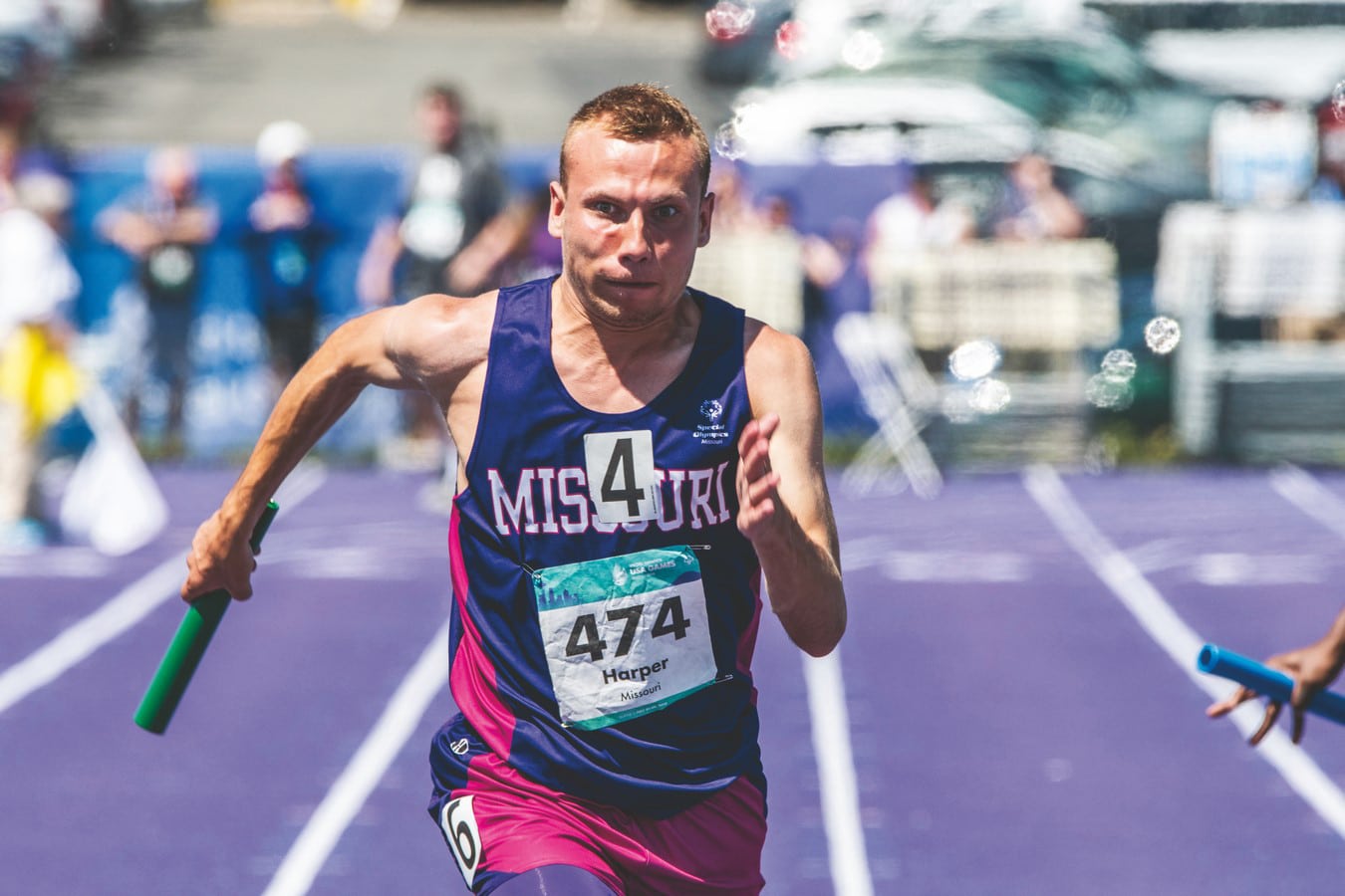 A track and field athlete in pink and purple Team Missouri attire runs toward the camera while holding a baton and eyes wide with a strained facial expression