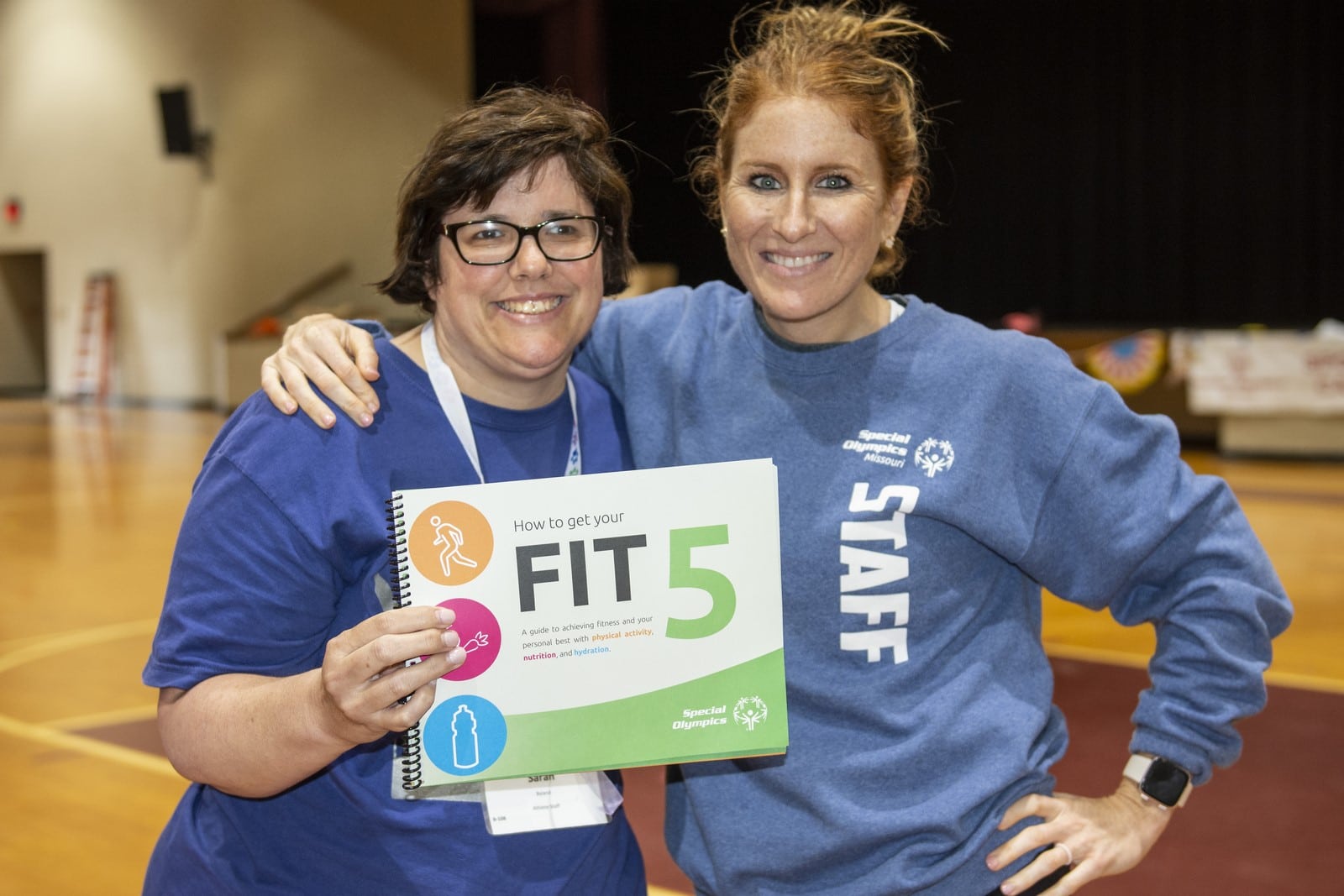 An athlete and staff member pose for a photo with a handout labeled "Fit 5" in their hand