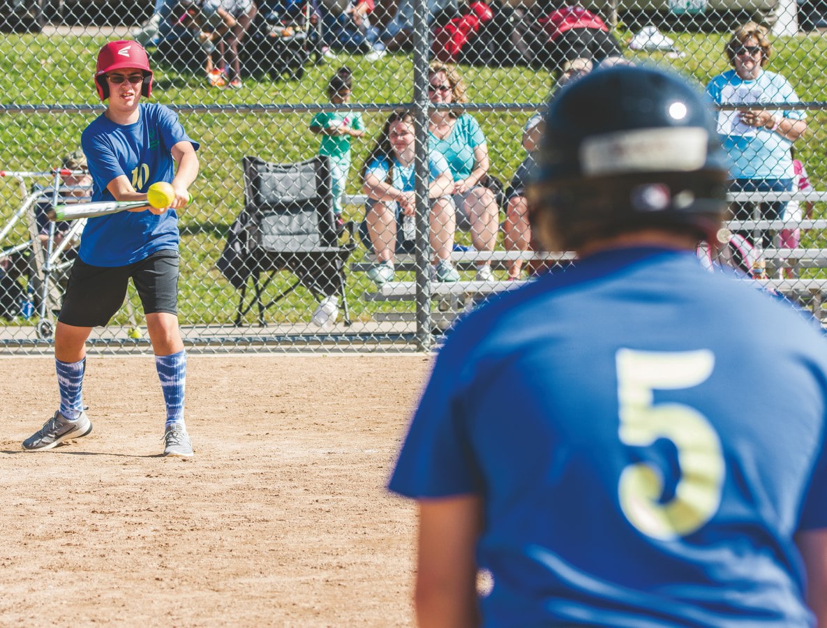 A softball player swings and strikes the ball while a teammate in the foreground stands at first base