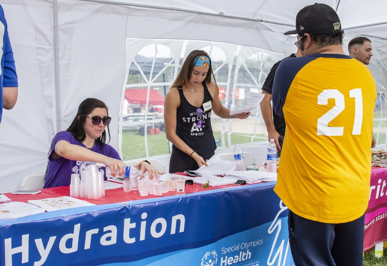 Two volunteers sit behind a hydration station and fill cups of water while an athlete stands and waits