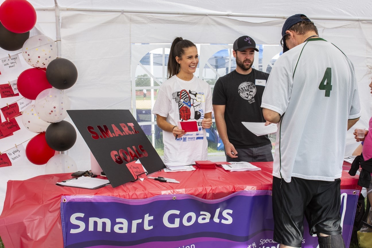 Volunteers stand behind a table labeled "Smart Goals" while an athlete writes on a card in front of them