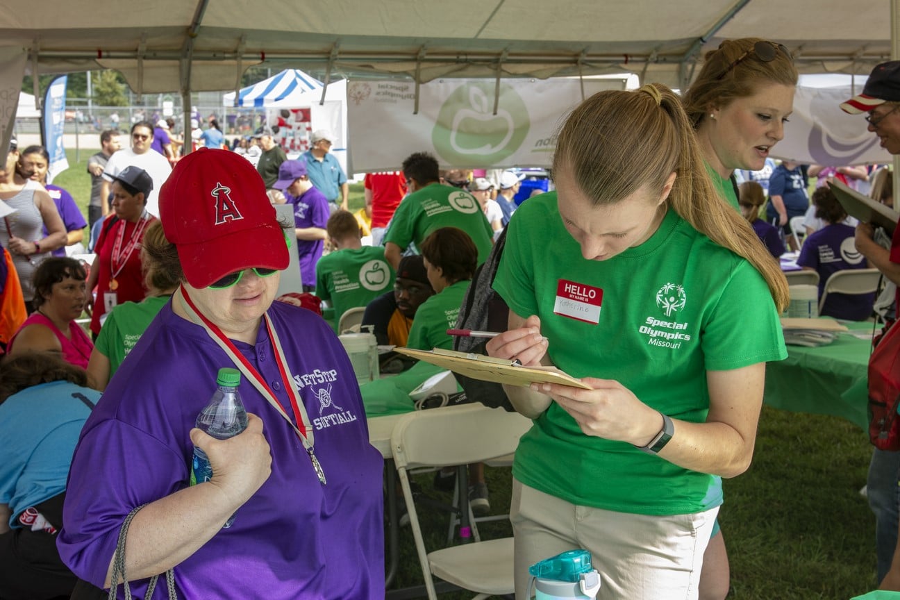 A health professional volunteer writes on a clipboard while an athlete stands near by