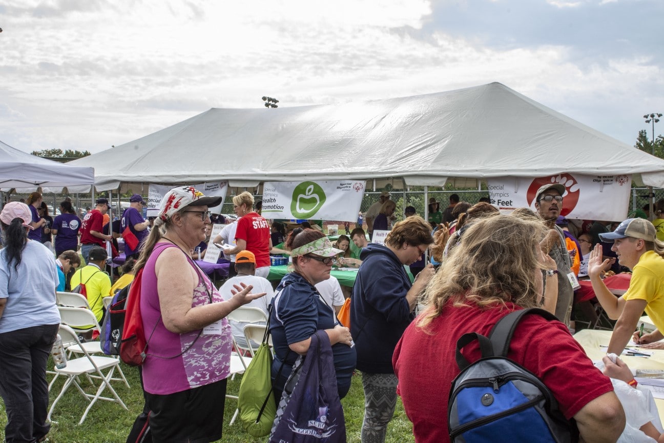 A group of athletes, coaches, and family members mill about around outdoor tents for Healthy Athletes