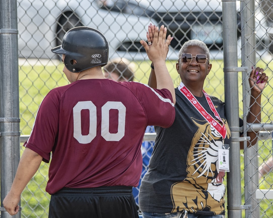 Smiling coach stands at dugout and high-fives athlete with softball helmet on