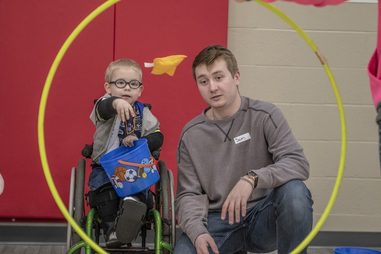 Young athlete in wheelchair holds blue bucket and tosses a yellow beanbag towards a yellow hula hoop while volunteer sits and watches