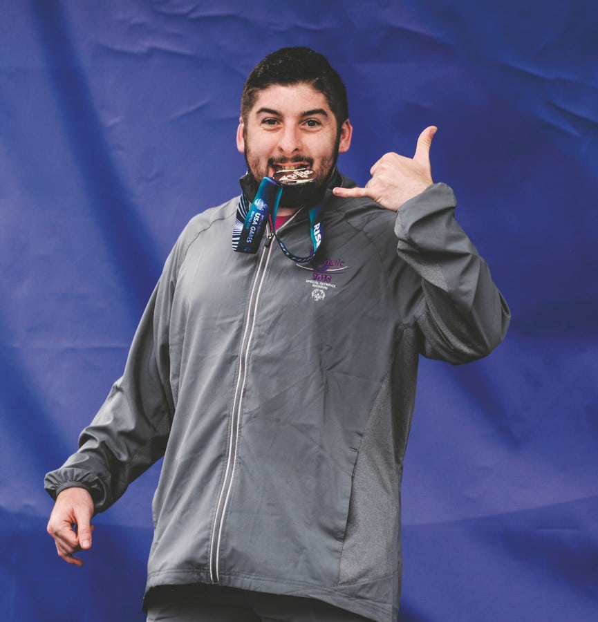 An athlete looks at the camera with a USA Games medal in their mouth while flashing a "hang loose" sign with their hands