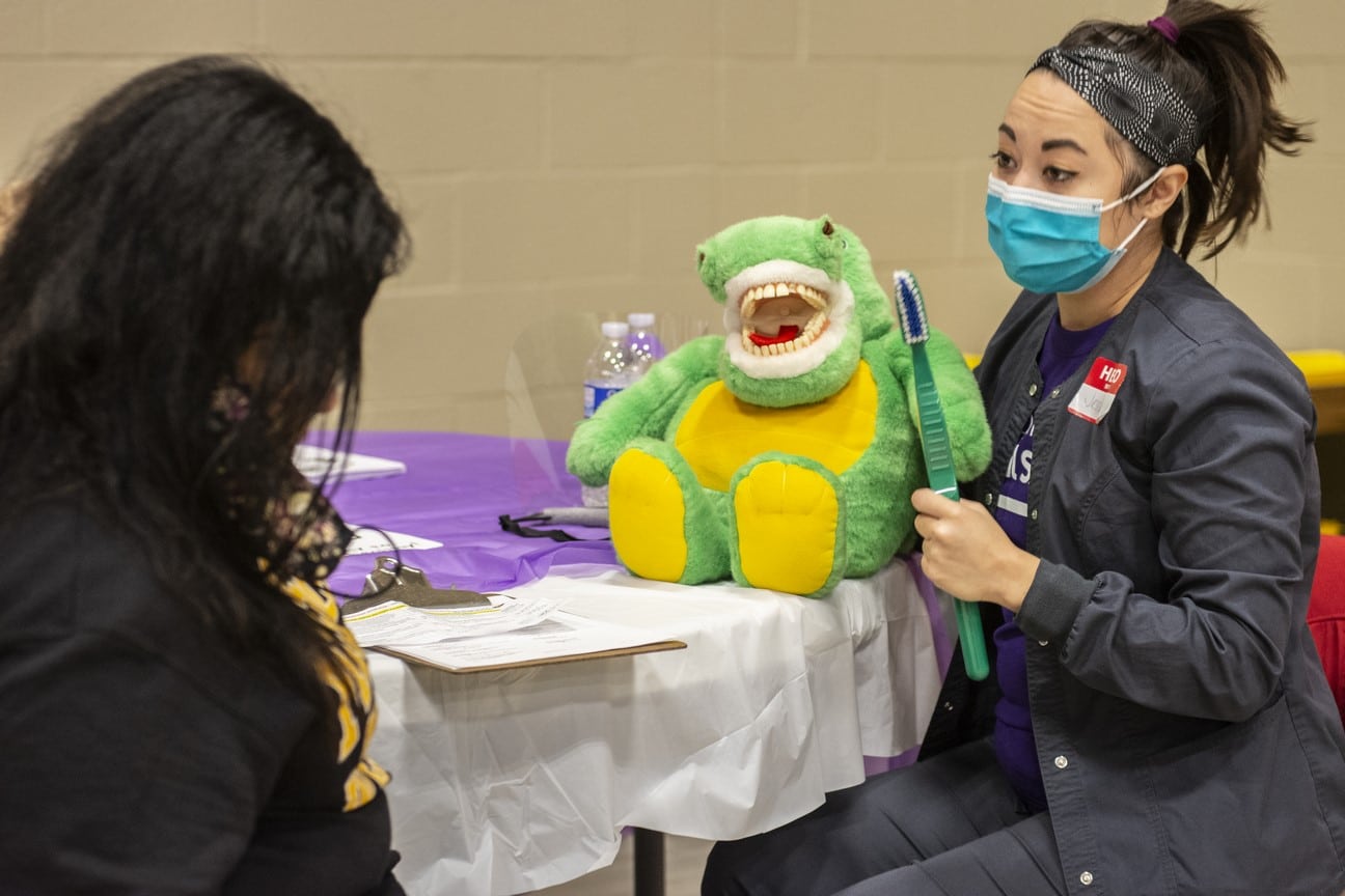 A health professional holds an oversized toothbrush and a stuffed animal and talks to an athlete about the proper way to brush their teeth
