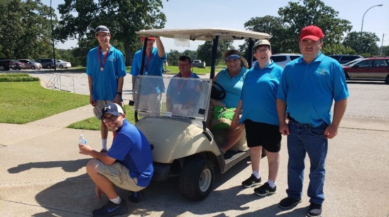 Athletes in bright blue shirts pose for a photo on the golf course in front of a golf cart