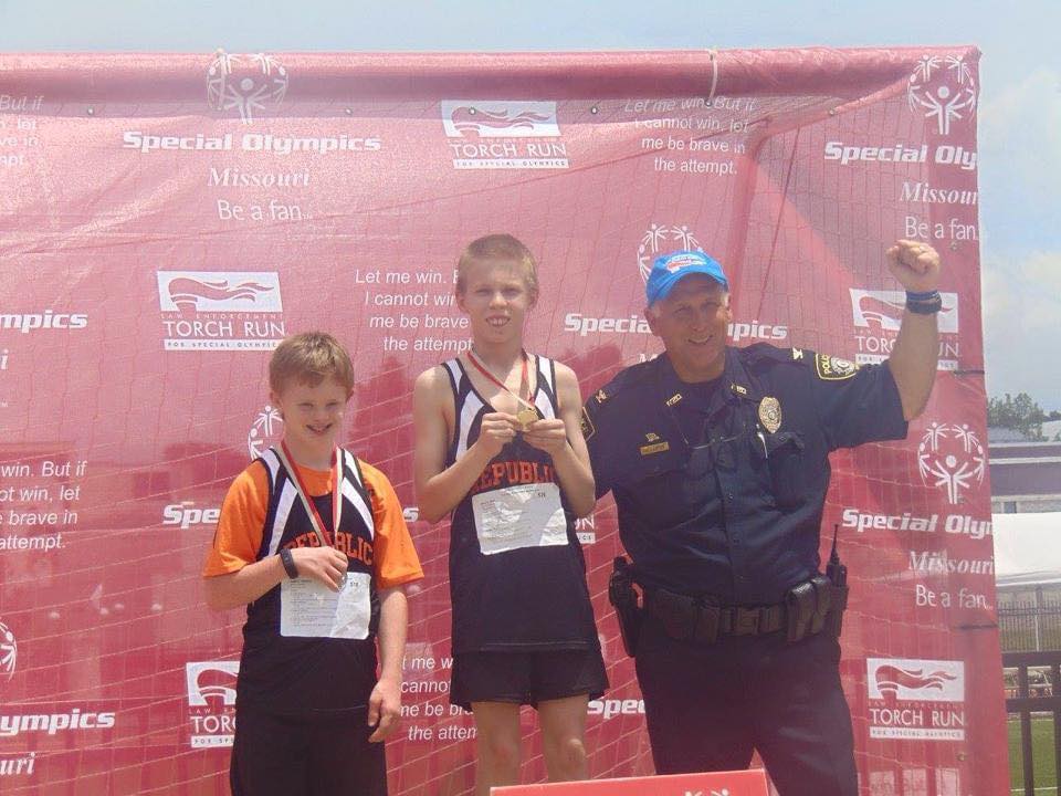 Two young athletes stand on a medal stand showing off their medals with a law enforcement officer raising their fist in the area and smiling