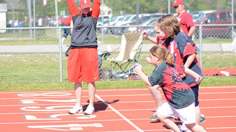 A group of athletes break off the starting line in track while a volunteer points a starter's pistol in the air