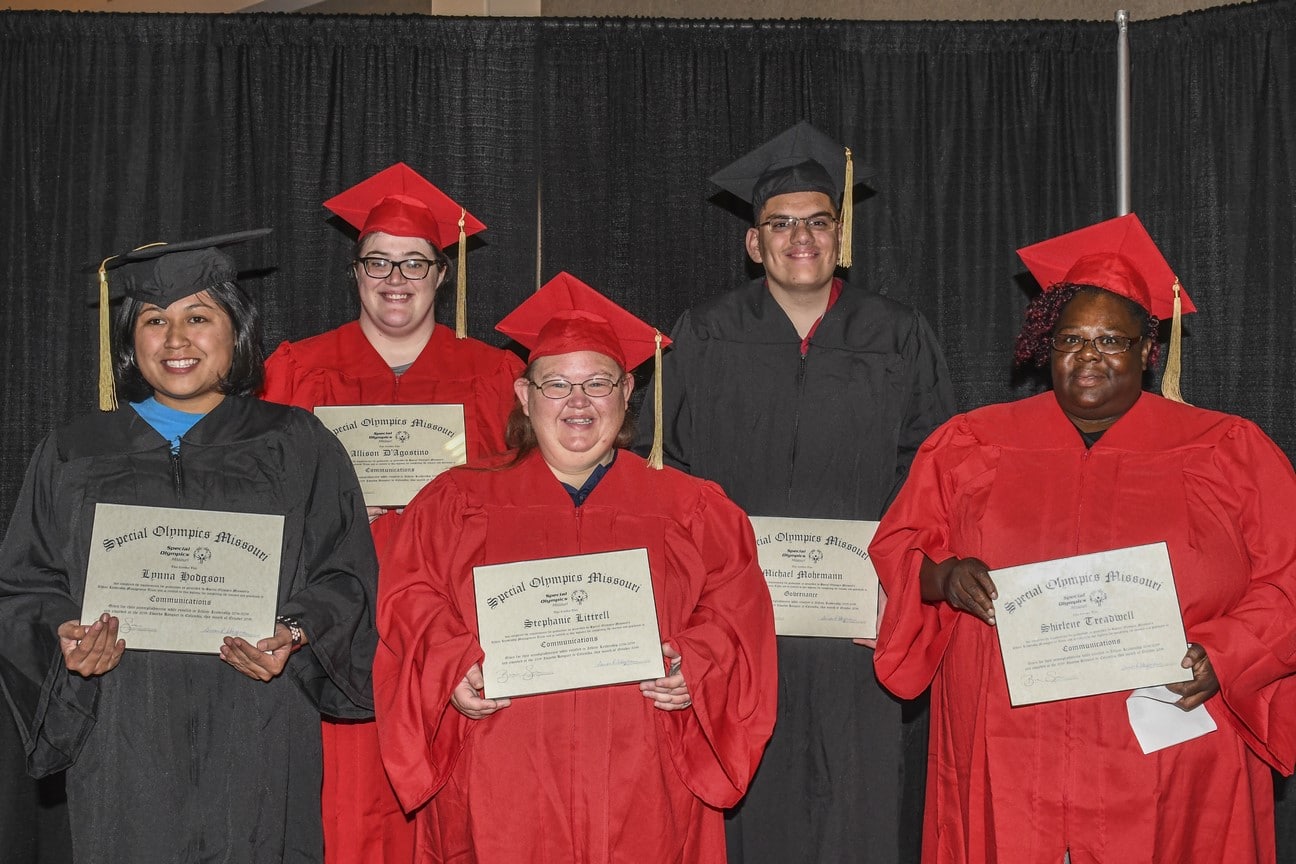 Five people wearing red and black graduation caps and gowns pose for a photo while holding framed degrees