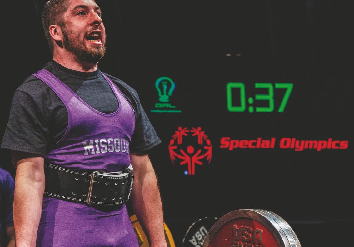 An athlete wearing a purple Team Missouri uniform successfully lifts a large amount of weight with a Special Olympics logo and countdown timer in the background