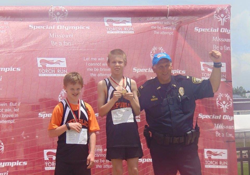 Republic Athletes Awards Two young athletes stand on a medal stand showing off their medals with a law enforcement officer raising their fist in the area and smiling