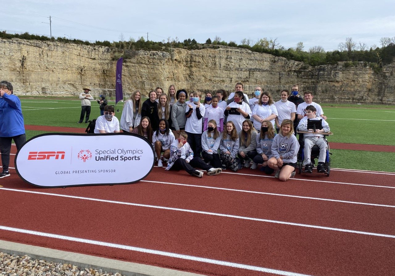 A group of athletes and volunteers pose for a photo on a track in front of a banner