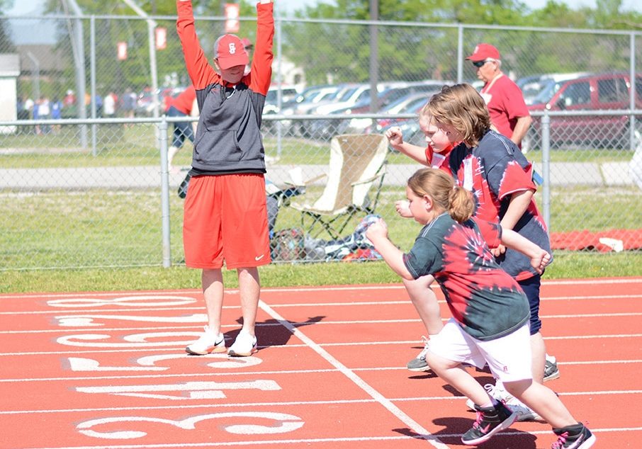 A group of athletes break off the starting line in track while a volunteer points a starter's pistol in the air