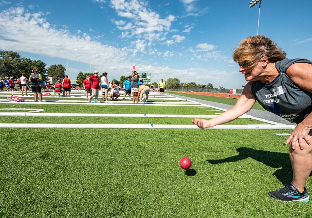 Bocce An athlete releases a bocce ball to roll down a turf field with many other bocce courts in the background